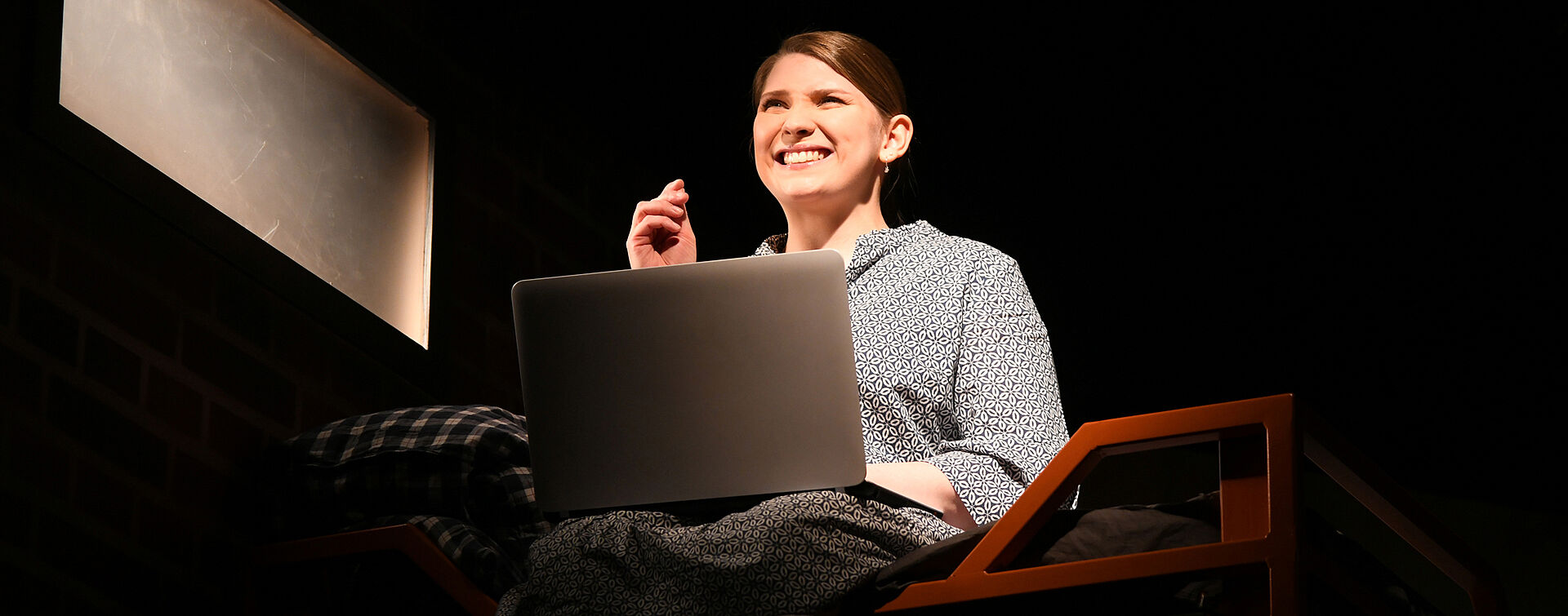 Lena Becker sitzt mit einem Laptop im Schoß auf einem Hochbett und hört Musik, dabei schnippst sie mit den Fingern. Sie trägt ein blau-weiß gemustertes Kleid.